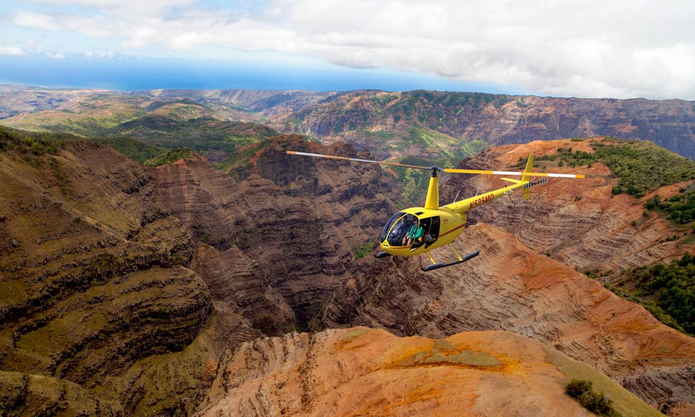 A yellow Robinson R44 helicopter flying above the dramatic ridges of Kauai’s Waimea Canyon